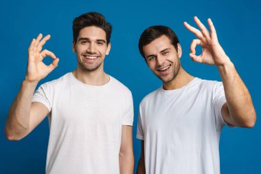 Neşeli yakışıklı adamlar gülümsüyor ve mavi arka planda iyi bir işaret gösteriyor.