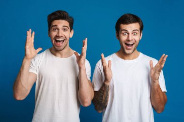 Excited unshaven two guys exclaiming with hands up isolated over blue background clipart