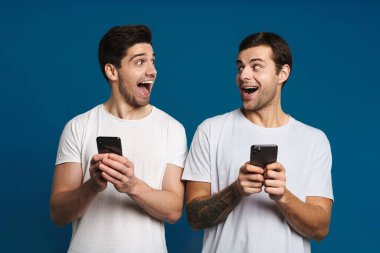 Mavi arka planda izole edilmiş akıllı telefonlarla poz verirken şaşkınlıklarını ifade eden iki adamı memnun ettim.