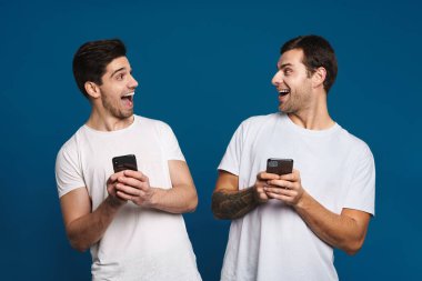 Mavi arka planda izole edilmiş akıllı telefonlarla poz verirken şaşkınlıklarını ifade eden iki adamı memnun ettim.