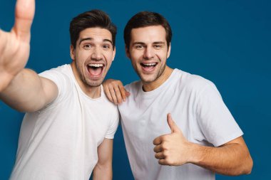 Mavi arka planda izole edilmiş selfie çekerken baş parmağını gösteren yakışıklı adamlar.