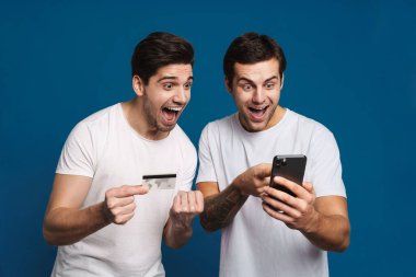 Delighted two guys posing with credit card and mobile phone isolated over blue background clipart