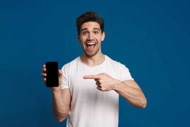 Mavi arka planda izole bir şekilde cep telefonunu gösteren ve işaret eden memnun bir adam.