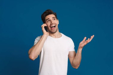 Mavi arka planda izole bir şekilde cep telefonuyla konuşurken gülümseyen mutlu traşsız adam.