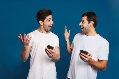 Mavi arka planda izole edilmiş akıllı telefonlarla poz verirken şaşkınlıklarını ifade eden iki adamı memnun ettim.