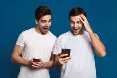 Mavi arka planda izole edilmiş akıllı telefonlarla poz verirken şaşkınlıklarını ifade eden iki adamı memnun ettim.