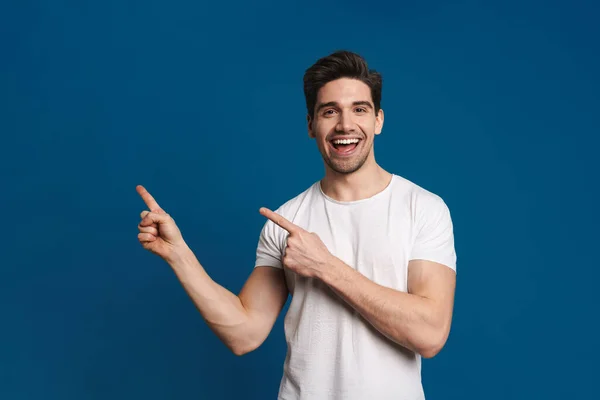 Happy unshaven guy smiling and pointing fingers aside isolated over blue background