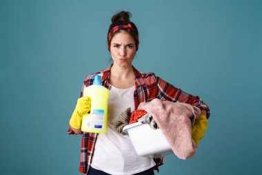 Önlüklü ve lastik eldivenli üzgün ev hanımı. Elinde temizleme aletleri var. Mavi üzerine izole edilmiş.