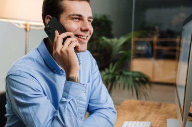 Gülümseyen genç girişimci modern bir ofiste otururken cep telefonu kullanıyor.