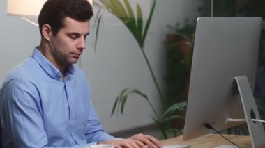 Gömlek giymiş, bardaktan bir şey içen ve ofiste bir masada otururken bilgisayarda yazan konsantre bir adamın yan görüntüsü.