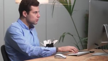 Gömlekli bir adamın, elinde sopayla bir kutudan bir şey yiyip, bir ofiste masada otururken klavyeye yazı yazmasının yan görüntüsü.
