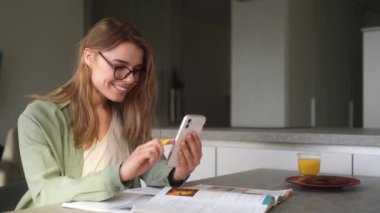 Gülümseyen kadın stüdyoda bir masada otururken telefon ekranından not defterine bir şey yazıyor.