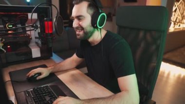 Kulaklık takan gülen bir adam evde oturan mikrofonla konuşurken profesyonel ve güçlü bir bilgisayardan çevrimiçi video oyunları izliyor.