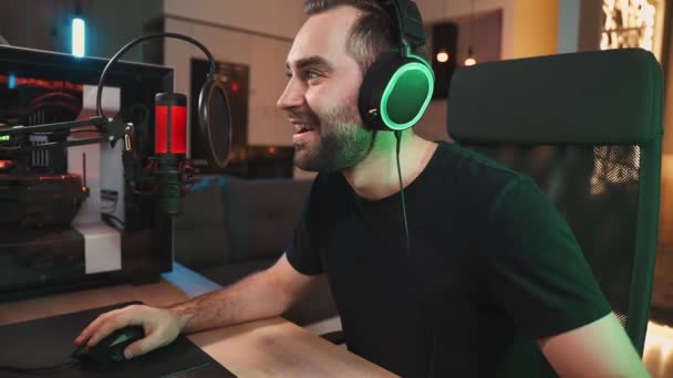 イヤホンを身に着けている笑顔の男のゲーマーは 自宅に座ってマイクに話をしながら プロの強力なコンピュータ上でオンラインビデオゲームをストリーミングしています — ストック動画