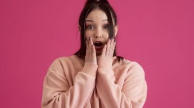 Şaşırmış genç bir kız kameraya bakıyor. Stüdyonun pembe duvarında tek başına duruyor.
