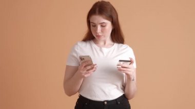 Memnun ve boyuna göre kızıl saçlı bir kadın kredi kartının bir kısmını akıllı telefonuna yazıp stüdyodaki bej bir duvarın arkasında duruyor.