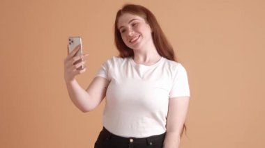 Yakışıklı ve iri yarı kızıl saçlı bir kadın stüdyodaki bej bir duvarın üzerinden izole edilmiş akıllı telefonunu kullanarak video bağlantısıyla konuşuyor.