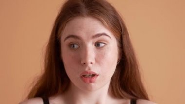 Kafası karışık ve boyunda kızıl saçlı bir kadının yakın çekim görüntüsü, stüdyodaki bej bir duvarın üzerinde tek başına duran omuzlarını silkiyor.