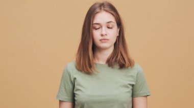 Kafası karışmış genç bir kadın, stüdyodaki bej bir duvarın arkasında durarak hızlı olmayan bir jest yapıyor.