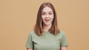 Gülümseyen genç bir kadın elini kaldırırken kameraya bakıyor. Stüdyodaki bej bir duvarın üzerinde tek başına duruyor.