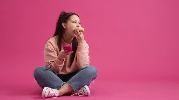 幸せな若い10代の女の子は彼女のスマートフォンを使用していますが ピンクのクリームとドーナツを食べながら スタジオのピンクの壁の上に隔離された床に座って — ストック動画