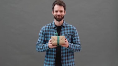 Ekose gömlekli mutlu bir adam gri bir stüdyoda dikilirken kameraya hediye veriyor.