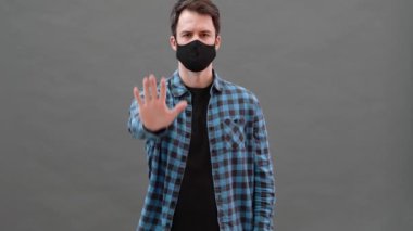 Tek kullanımlık koruyucu maskeli kendine güvenen bir adam gri bir stüdyoda bir el hareketi gösteriyor.