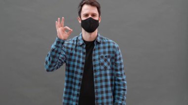 Siyah koruyucu maskeli mutlu bir adam gri bir stüdyoda dururken iyi niyet gösterisi yapıyor.