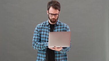 Gözlüklü gülümseyen adam dizüstü bilgisayarda bir şey arıyor ve gri bir stüdyoda dururken elinde tutuyor.
