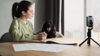 Gülümseyen kadın odada masada otururken telefon kamerasında köpeğini okşuyor.