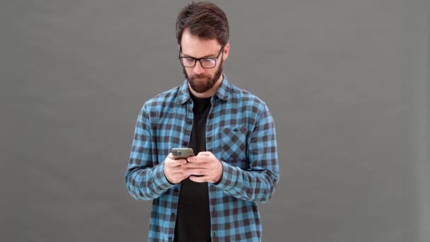 一个微笑的男人 站在灰色的工作室里 戴着眼镜 在电话里打字 — 图库视频影像