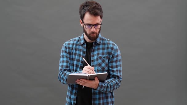 灰色のスタジオに立っている間にノートに何を書くべきかを考えている眼鏡をかけた瞑想的な男 — ストック動画