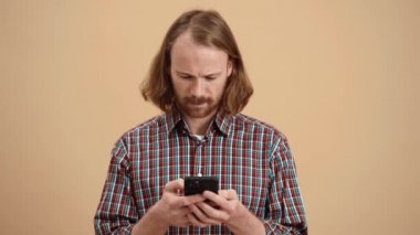 Ekose gömlekli, uzun saçlı gülümseyen adam bej renkli bir stüdyoda dikilirken kameraya bakıyor.