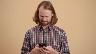 Ekose gömlekli, şok olmuş adam telefona bakıyor ve bej renkli bir stüdyoda dururken yüzünü buruşturuyor.