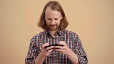 Uzun saçlı, ekose gömlekli mutlu adam telefonda oyun oynuyor ve bej renkli bir stüdyoda durup zaferle seviniyor.