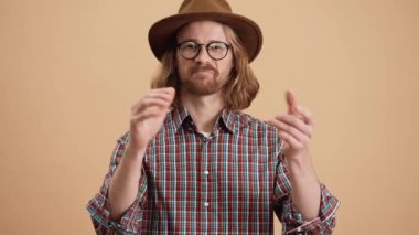 Şapkalı ve gözlüklü gülümseyen bir adam el çırpıyor ve bej renkli bir stüdyoda dikilirken iki parmağını kaldırıyor.