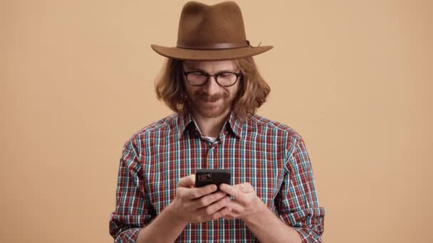 Şapkalı Gözlüklü Gülümseyen Adam Bej Bir Stüdyoda Dururken Telefona Bakıyor — Stok video