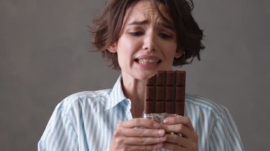 Kısa saçlı, elinde çikolatayla ikinci dilimi yemek isteyip gri bir stüdyoda reddeden üzgün kadın.