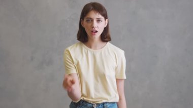 Gri stüdyoda dikilirken işaret parmağıyla sessizce hareket eden gülümseyen kız.