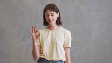 Kısa saçlı gülümseyen kız gri stüdyoda dururken iyi niyet gösterisi yapıyor.