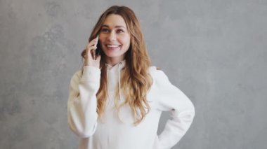 Kapüşonlu mutlu kadın gri bir stüdyoda dikilirken telefonla konuşuyor.
