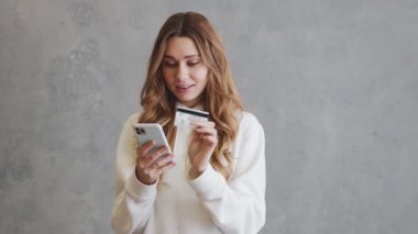 Gri bir stüdyoda dururken banka kartı numarasını yazan kapüşonlu olumlu kadın.