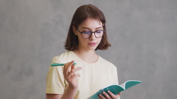 グレーのスタジオに立っている間にノートに何かを書く眼鏡の普及した女の子 — ストック動画