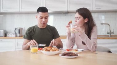 Gülümseyen genç çift beyaz mutfakta otururken kahvaltı yapıyor.
