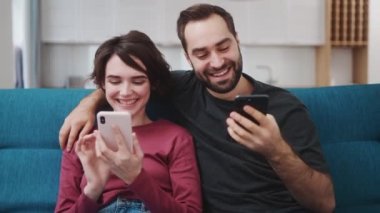 Gülümseyen çift, evdeki kanepede otururken akıllı telefonlarını kullanıyor.