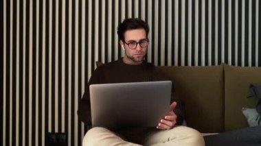 Yoğunlaşmış bir adam yatakta oturmuş çizgili odada dizüstü bilgisayara bakıyor.