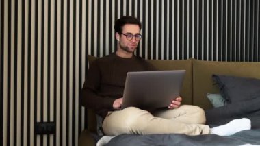 Yatakta oturmuş çizgili odada dizüstü bilgisayara bakan gülümseyen bir adam.