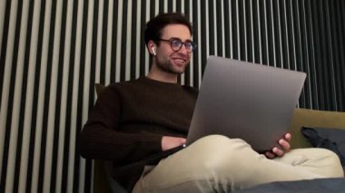 Çizgili odada dizüstü bilgisayara bakarken konuşan ve yatakta oturan gülümseyen bir adam.