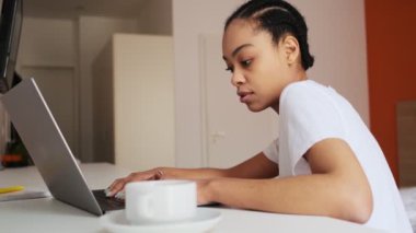 Düşüncelere dalmış Afrikalı kadının turuncu odada elinde bir bardakla dizüstü bilgisayarına bir şey yazışının yan görüntüsü.