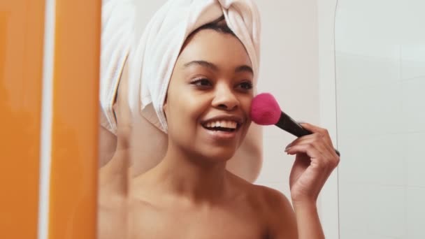 非洲妇女穿着毛巾在浴室的镜子中看着自己 用刷子擦拭自己的脸的特写镜头 — 图库视频影像